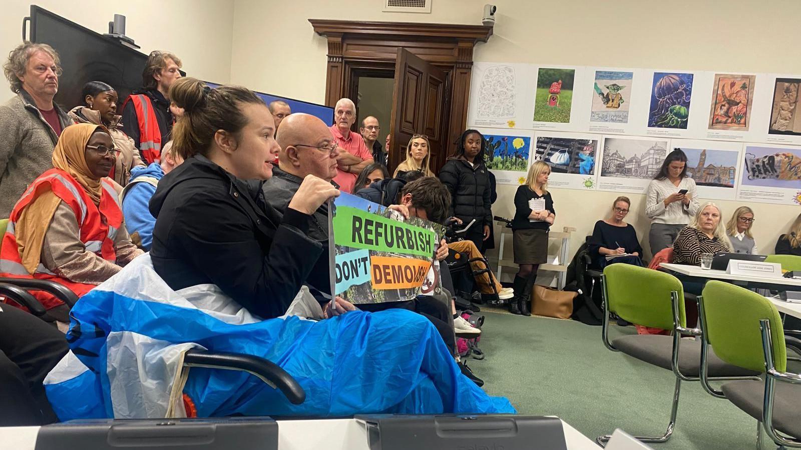 A group of people are sitting in a room holding up a sign that reads "refurbish don't demolish"