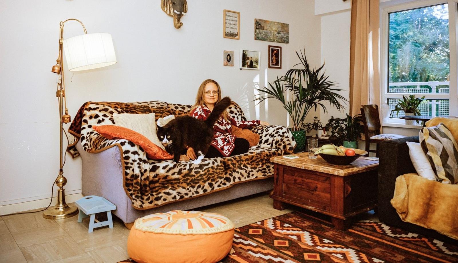 A lady sat on a sofa in her living room with her cat. The sofa has a leopard print throw on it and there are some pictures on the wall in the corner and a window to her right.