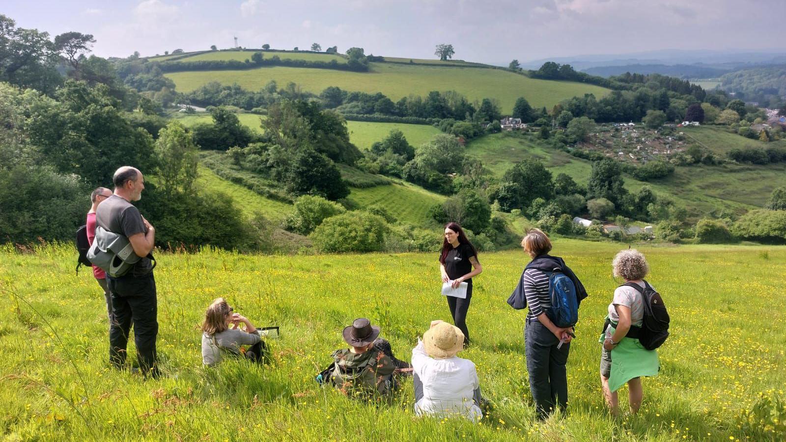 Claire Inglis is talking to a group of seven visitors to the Bowden Pillars site. Three of the visitors are sitting in the field, the others are standing. The field is lush with grass and covered with buttercups. There are rolling hills in the distance covered with trees and hedging. There are also a few homes dotted through the landscape.