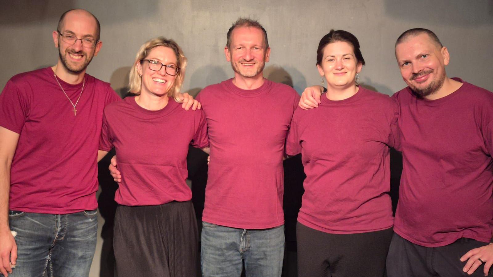 A group of five people, four men and one woman, stand in a line with arms linked as they smile at the camera. They are all wearing matching red T-shirts
