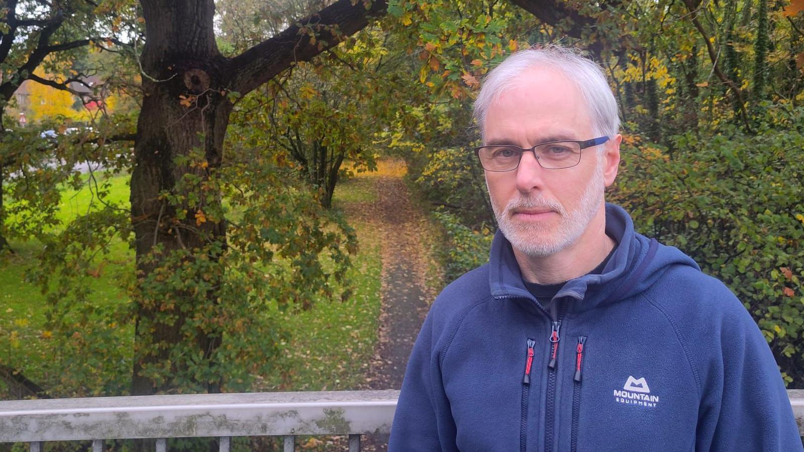 Adrian Howlett (white hair, beard, glasses) in front of a background of path and trees