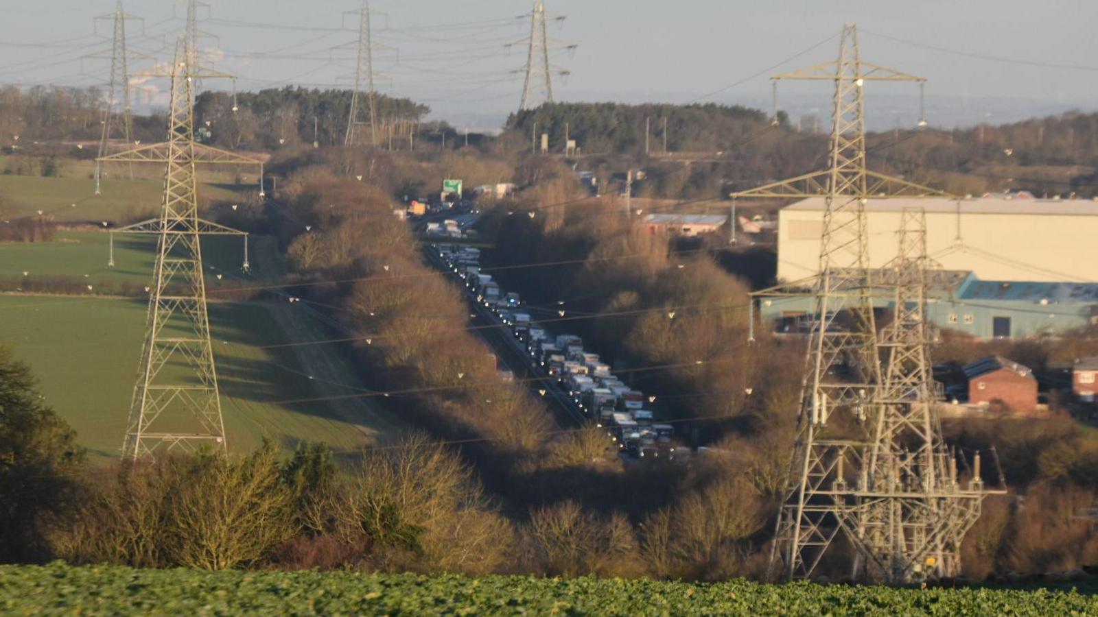 A aerial view of the A1 close to the scene of a crash which killed a baby boy. A long queue of traffic is seen backing up into the distance. The carriageway is surrounded by large trees and bushes. Green fields and electrical pylons are also visible.