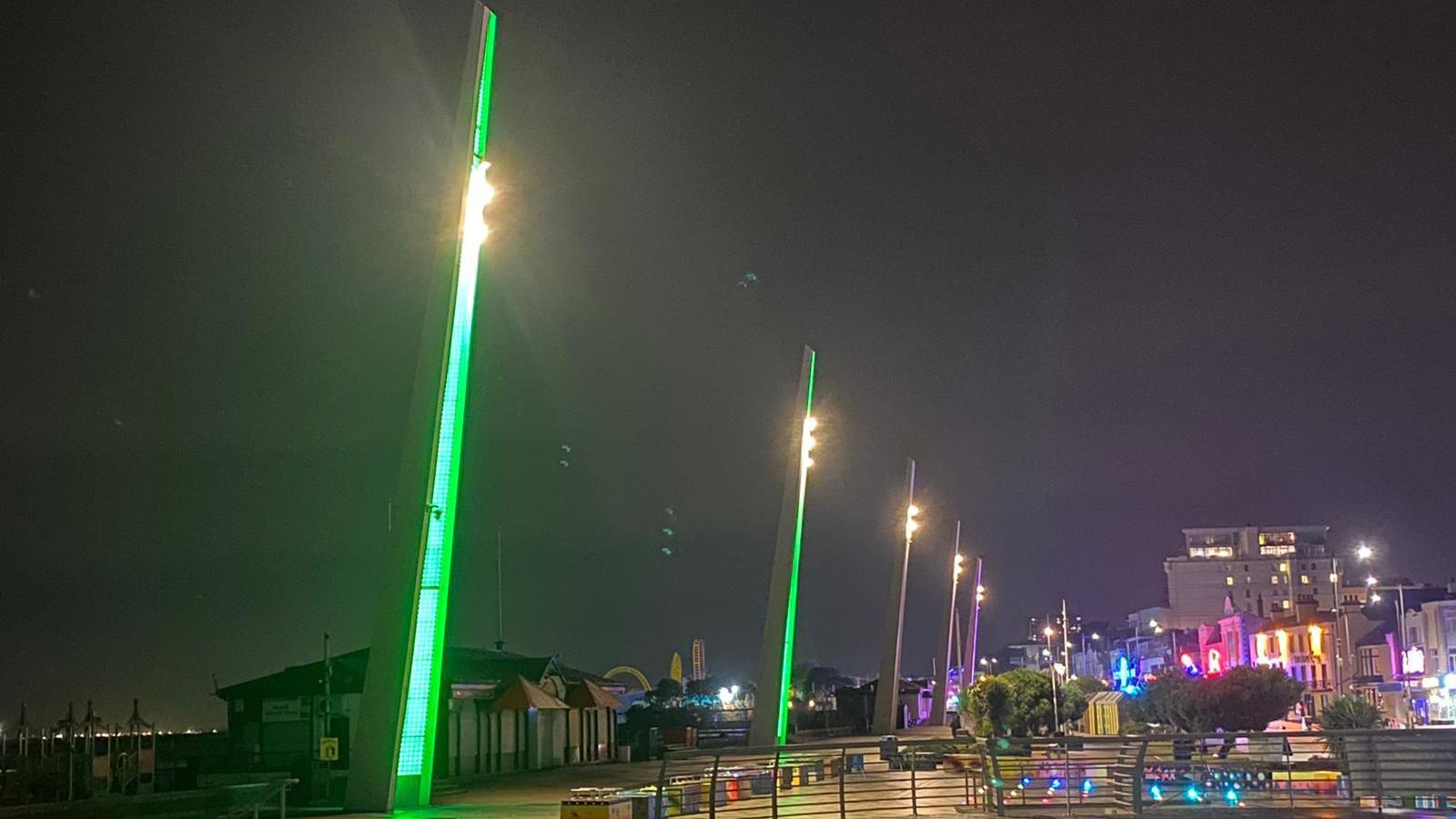 Large pillars next to the beach in Southend-on-Sea are illuminated green. In the background there are various buildings on the seafront.