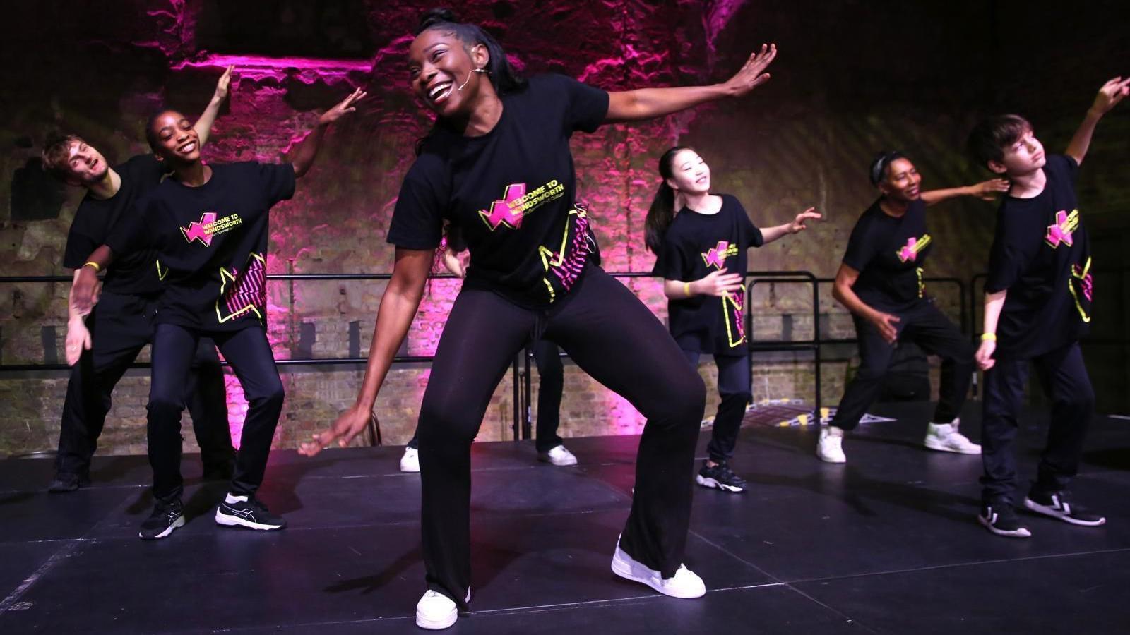 A group of young dancers, wearing black yellow and pink tops with black trousers, perform on stage