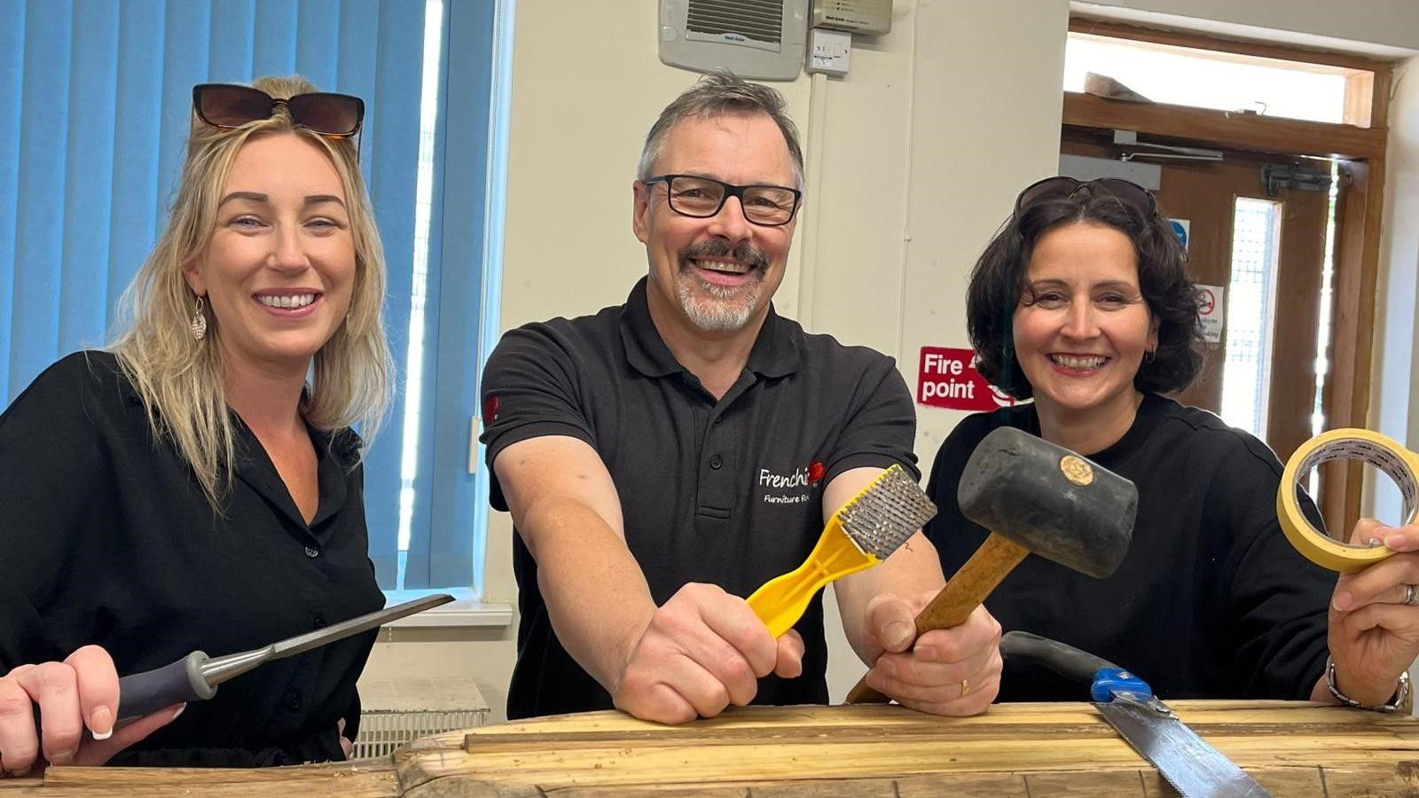 Emma Freemantle, Clerk of Wroughton Parish Council, Kev Prosser of Renew Swindon and Amanda Woodhead stood at a wooden bench holding tools. They are all looking into the camera and smiling.