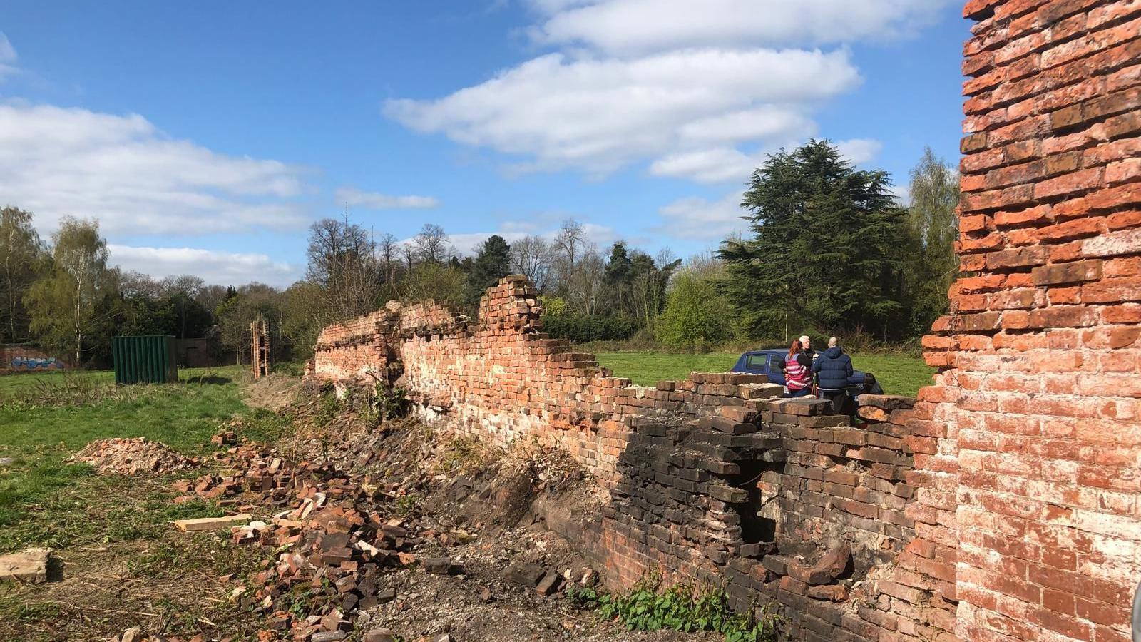 A partly collapsed red brick wall, extending through park land