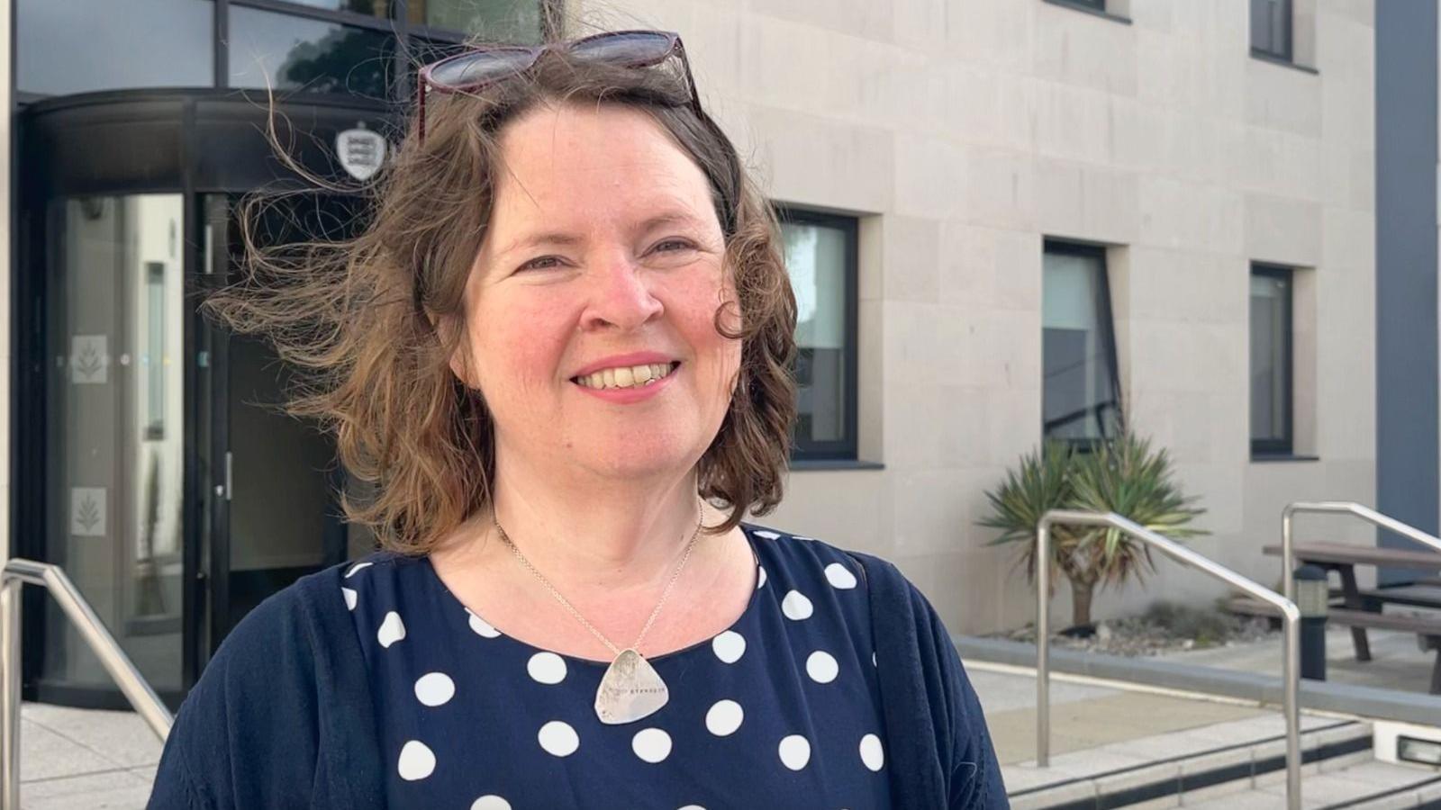 Daniele Harford-Fox - a woman with brown shoulder length curly hair, smiling with sunglasses on her head, wearing a navy cardigan and a navy top with white spots on it, a silver necklace in the shape of a soft angled triangle, background of a grey building with stairs and a table benches in front to the right