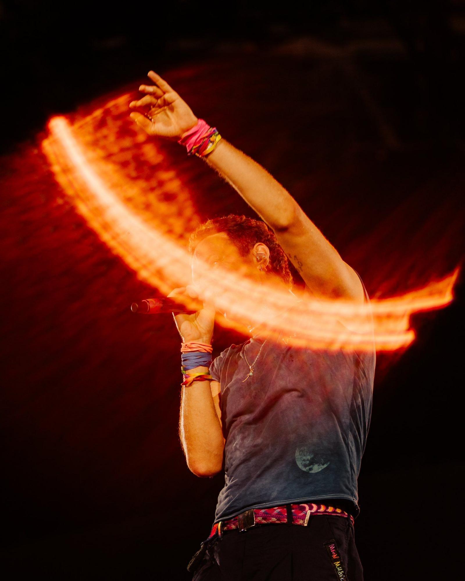Chris Martin is seen singing, with his face obscured by a streak of light