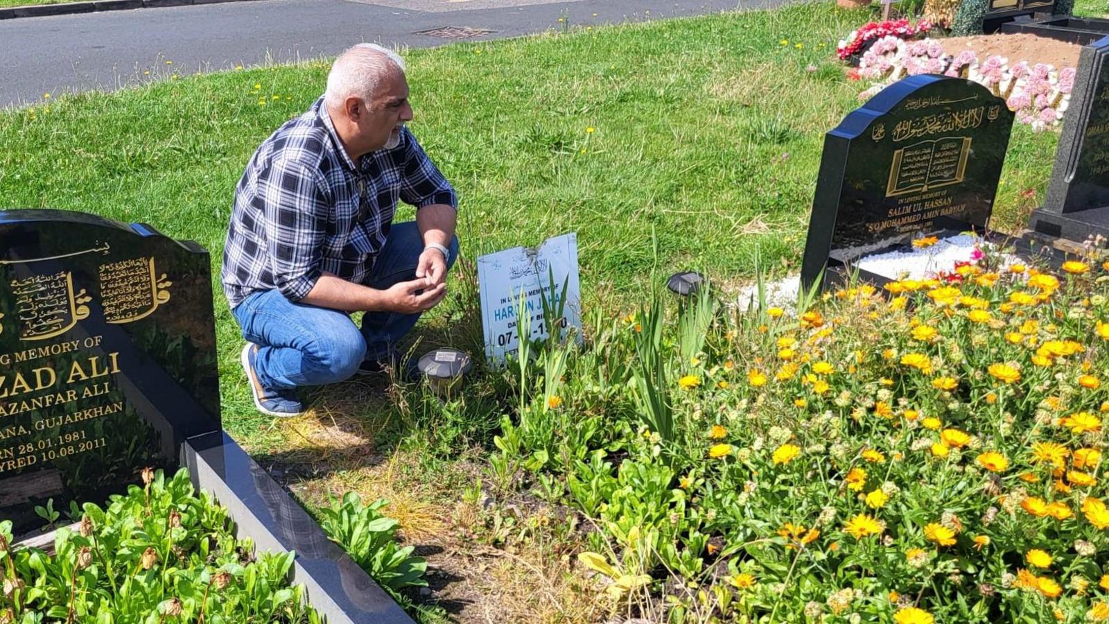 Tariq Jahan crouches at his son's grave. He is dressed in a checked shirt and jeans, with a number of graves visible, including his son's, planted with yellow flowers.