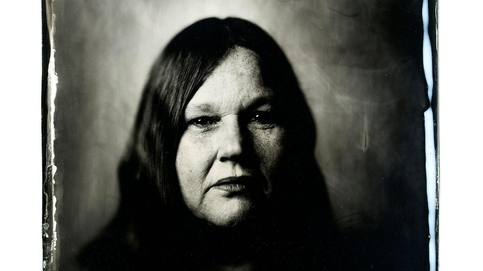 A black and white portrait image of Lorraine Maylan, taken as part of the project. She has long dark hair parted in the middle and is unsmiling as she looks into the camera.