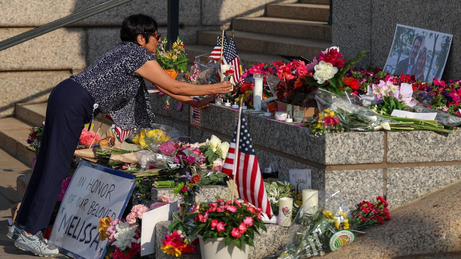 A display of flags and flowers fills the image as a woman leans over to lay a flower in the middle of the arrangement