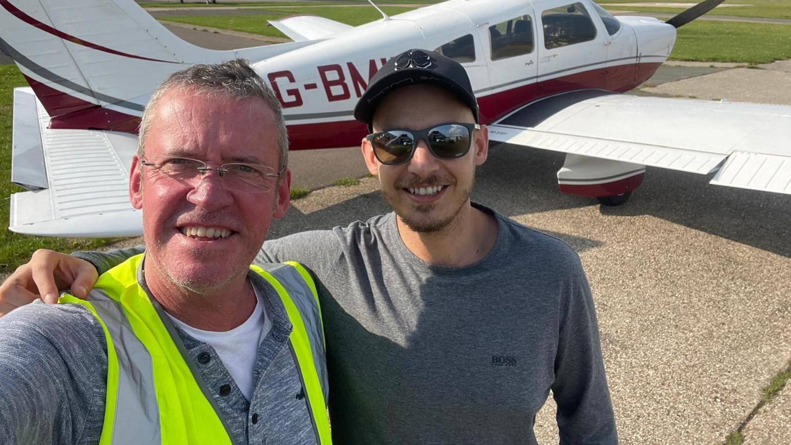 An older man (left) and a younger man wearing a cap and shades in front of a light aircraft on a runway