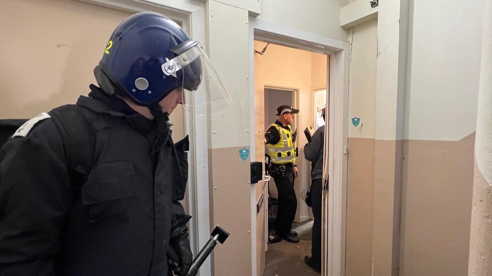 A police officer with a battering device stands outside the broken front door of a flat. Other police officers are visible within the property.