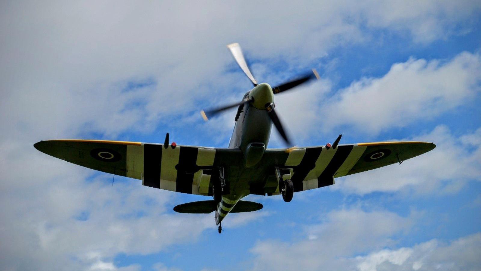 A spitfire which is light green and has black and white stripes on the wings flying against a blue sky with patches of white cloud. The propeller at the front of the plane is blurred as it is moving.