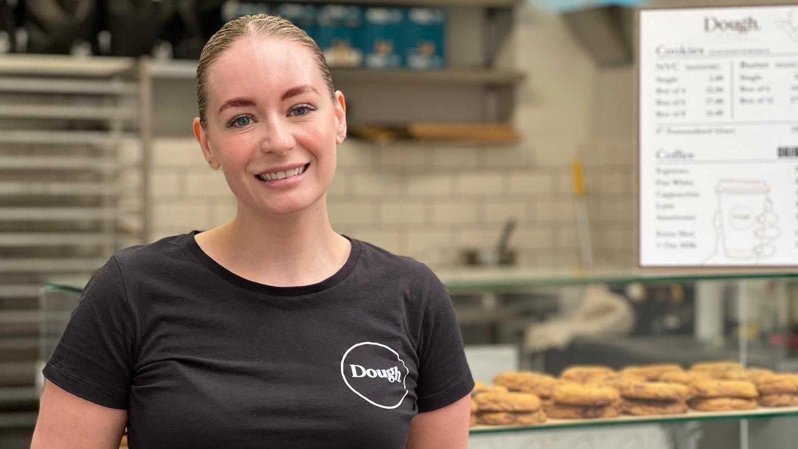 Ruby Price - a young woman smiling with slick back light brown hair, light makeup wearing a black shirt with a circle logo on her right chest with the word Dough in it, background of a bakery with a menu sign of cookie prices and coffee options and prices