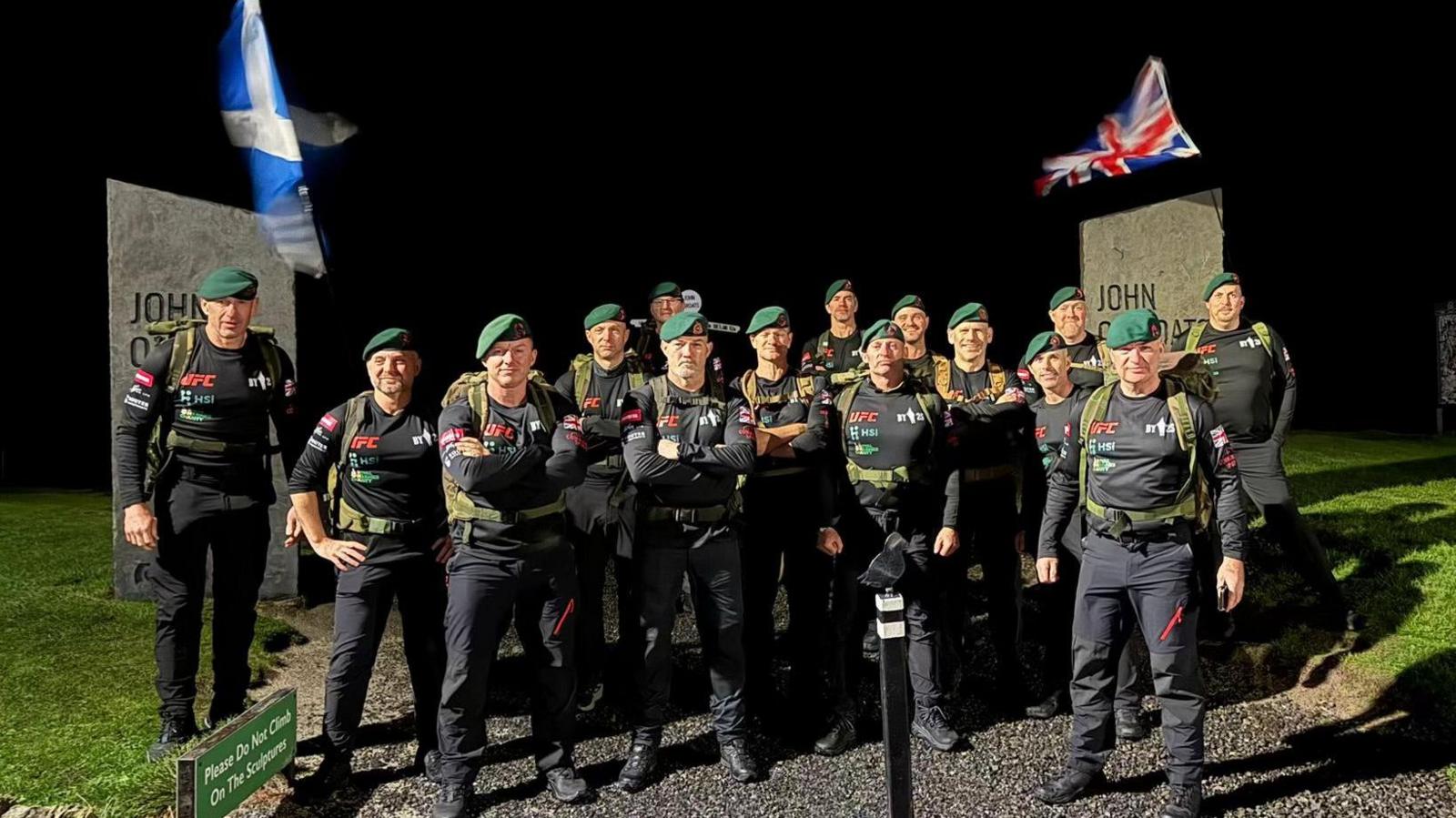 A group of men in black jumpers and trousers and green berets. Some of the men have their arms folded and they are looking into the camera on a dark night.