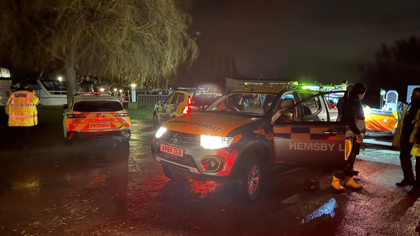 Emergency service vehicles, many with their lights on, parked by what appears to be a marina. Boats can be seen in the background