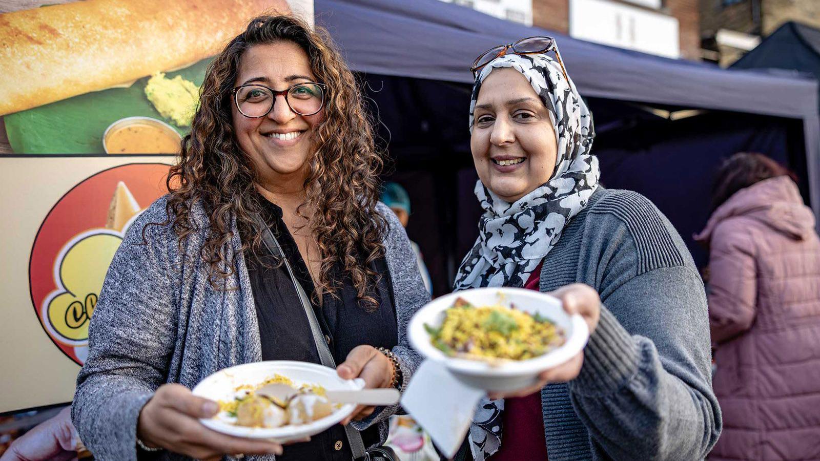 Two women show the camera plates of food. The woman on the left has long curly brown hair and the woman on the right is wearing a black and white head scarf. They are standing outside a food tent.