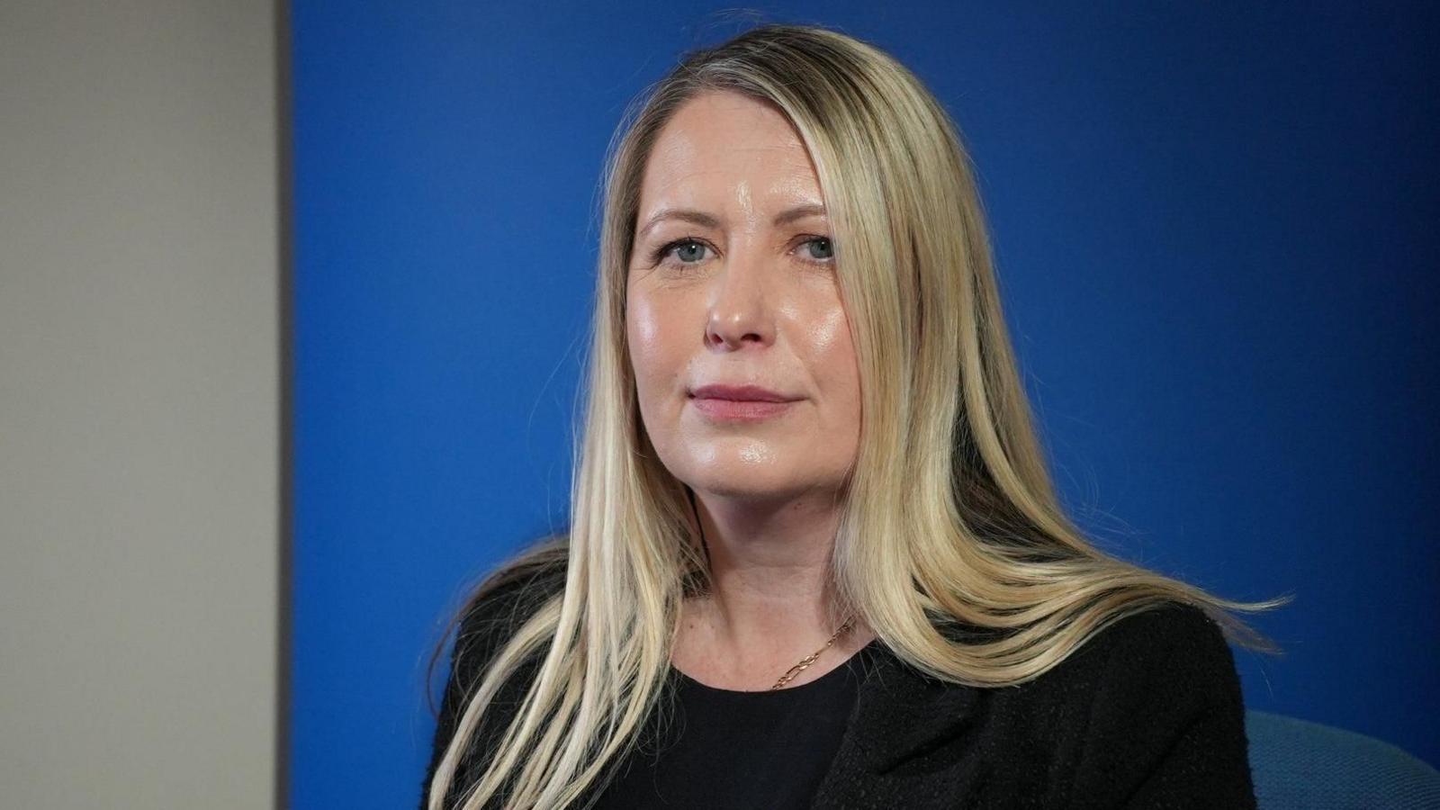 Leanne pictured looking into the camera. She's wearing a black blazer with a black top on underneath. She's sat in front of a royal blue backdrop with a white wall visible beyond it.