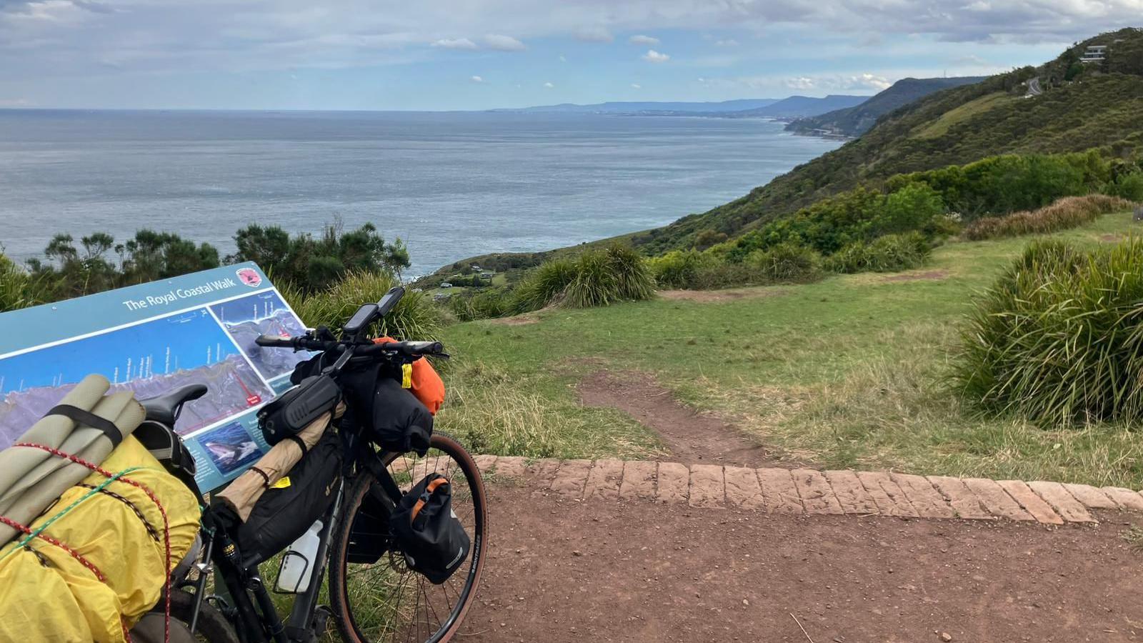 A pushbike laden leans against a sign reading: "The Royal Coastal Walk." In front of it is a stretch of green coastline.