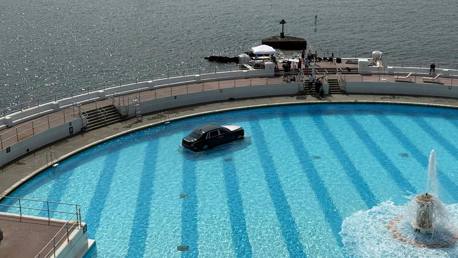 A Rolls Royce car on a submerged plinth in Tinside Lido in Plymouth on a sunny day. A fountain in the middle of the pool is spouting out water. The car is mostly black with a silver bonnet. A film crew is gathered on the outside edge of the pool.