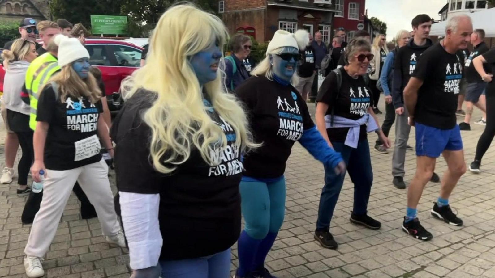 People dressed in blue trousers and wearing blue facepaint doing a warm-up exercise in the street.