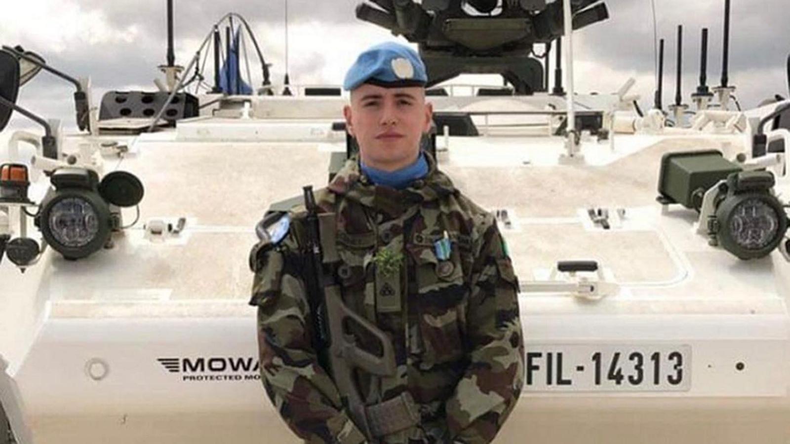 Irish soldier Seán Rooney, wearing camouflage clothes and a blue beret. He is holding a gun and standing in front of an armoured vehicle.