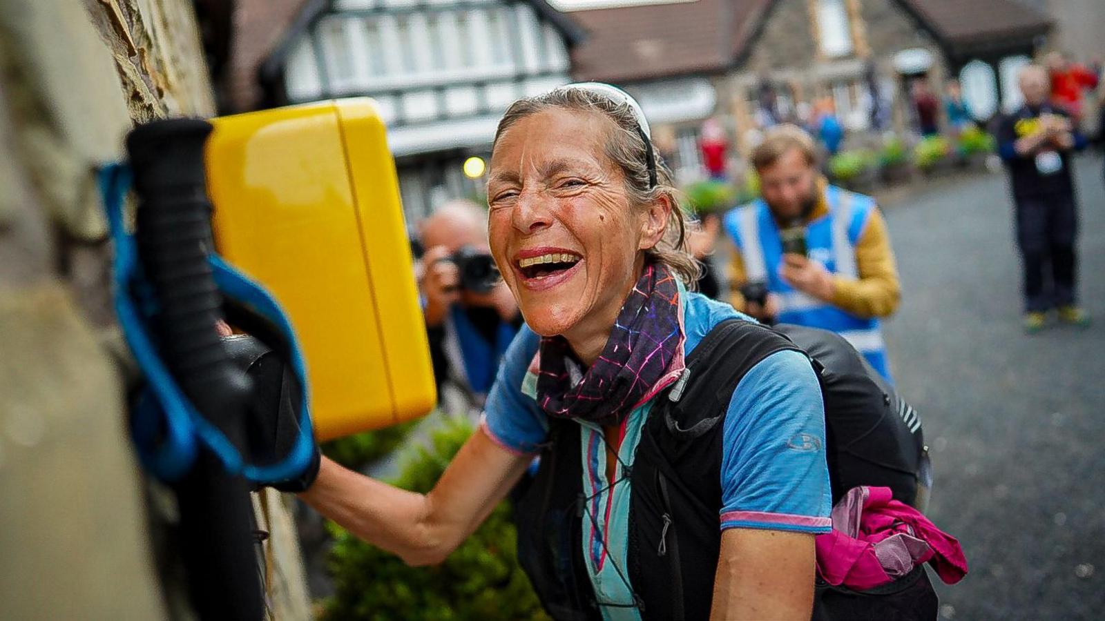 Anna Troup, who is kitted out in trail running attire, including shorts, running rucksack, poles, sunglasses and a sports watch, smiles as she reaches a rest point during the Summer Spine Race. She smiles as she stops in front of race marshals.