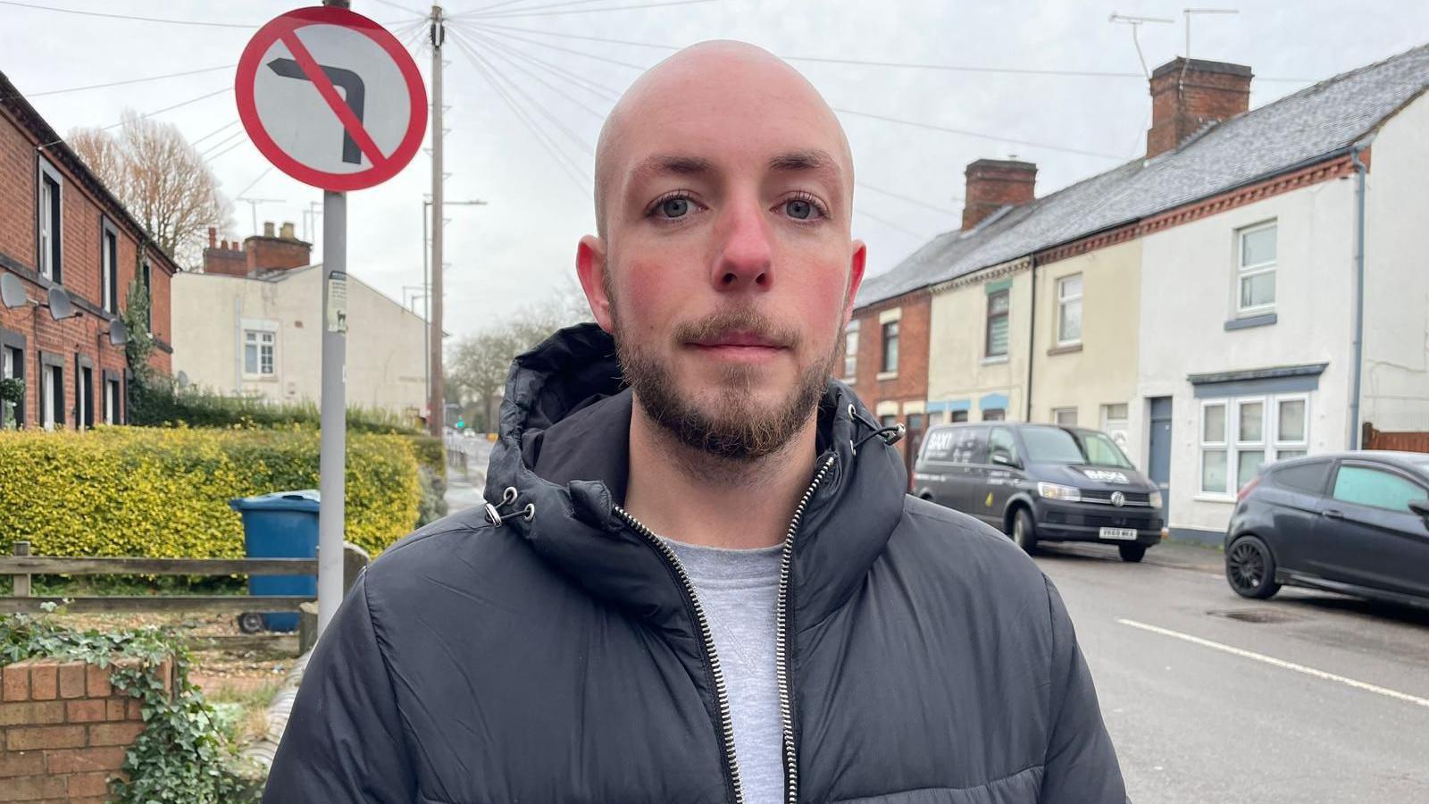 A young man with a shaved head and a short beard stands on a residential street with cars parked along the side of the road. He is wearing a grey top under a black hooded jacket.