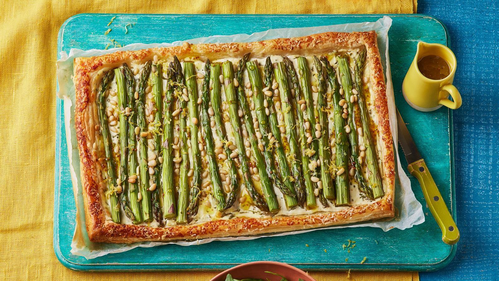 A puff pastry asparagus tart, topped with pine nuts on a painted blue wooden board with a sharp knife. 