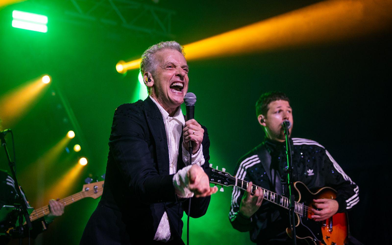 Peter Capaldi - a man with short grey hair wearing a black suit jacket and white shirt singing into a microphone with a guitarist behind him