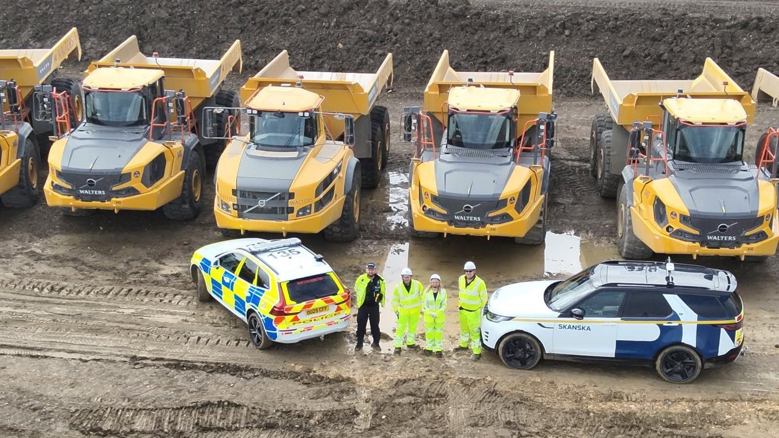 An aerial view of five large yellow vehicles. In front are four people in high vis. Next to them is a police car.