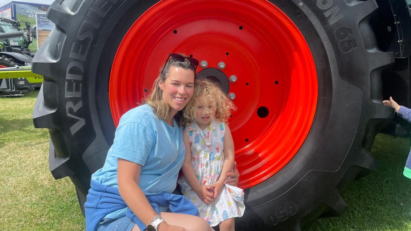 A young girl sitting in a red and black large tractor wheel. The girl has curly blonde hair and is wearing a ice cream themed multicolour dress. Her mother is stood next to her and is wearing a blue t shirt and shorts. She has brown hair and sunglasses on top of her head.  