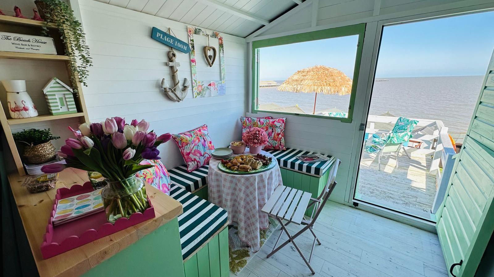 The view outside the beach hut, which looks at the sea. Inside the hut is a table which is surrounded by a bench seat. It has food on top. The bench seat has a black and white-striped pillow. There are also pink pillows.