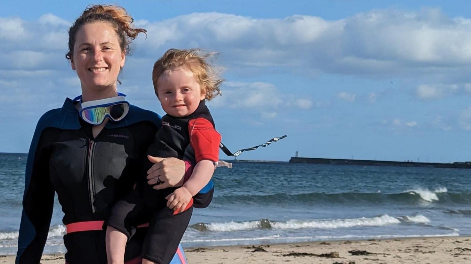 A smiling woman in a wetsuit holding a toddler