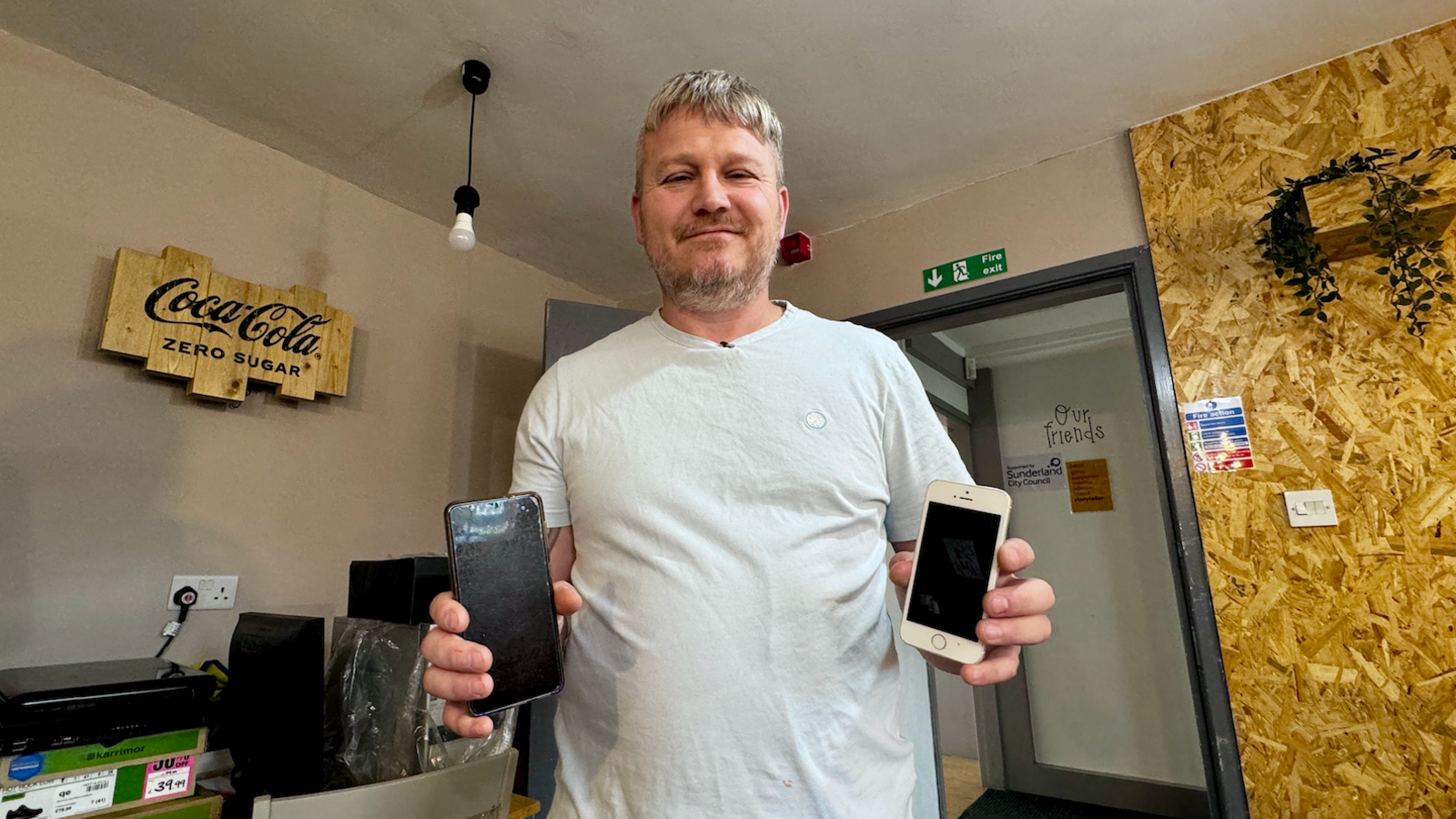 A man with short light brown hair holds up two donated mobile phones. He is wearing a light blue-coloured T-shirt.