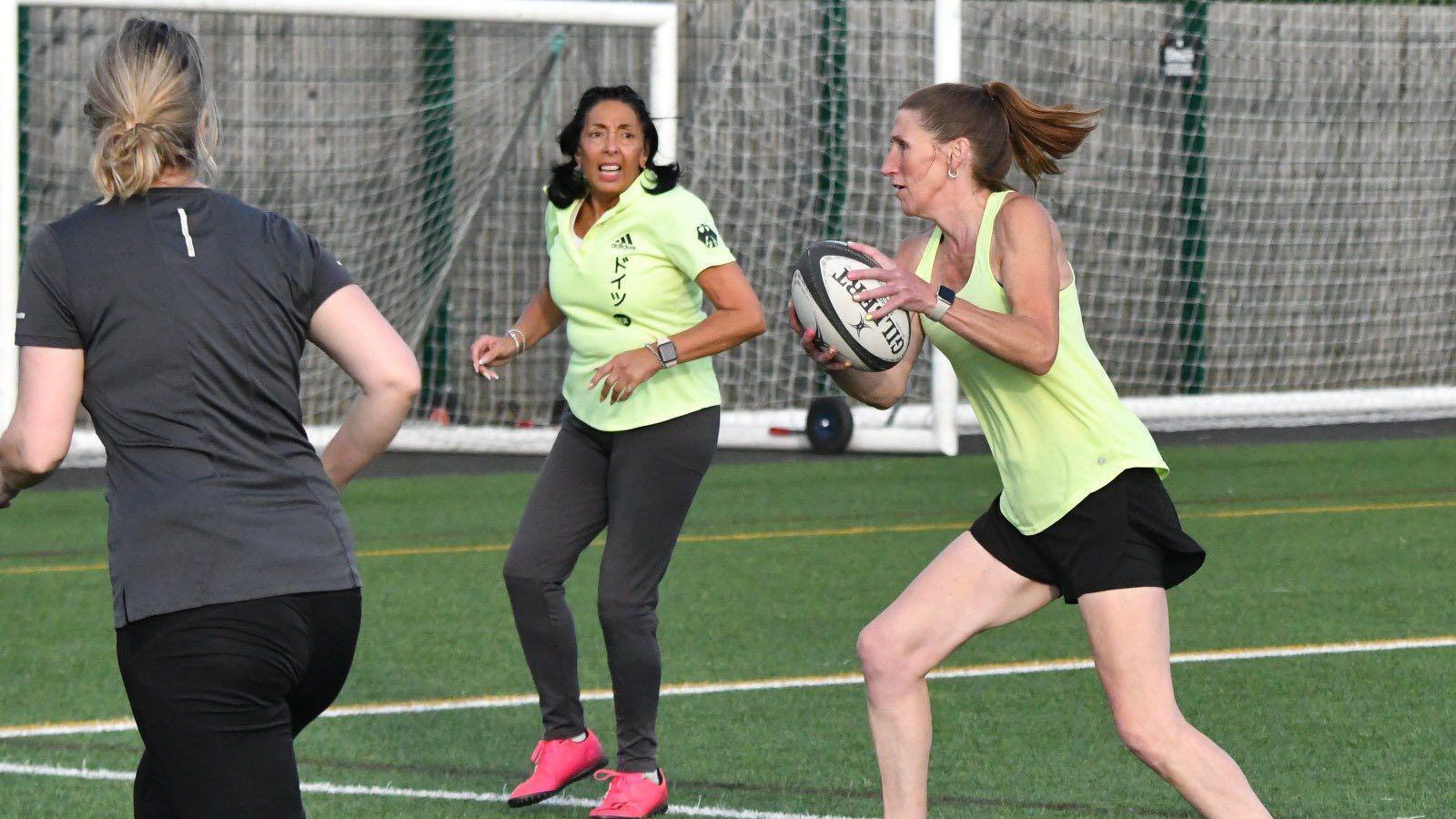 Three women playing touch rugby. A slender woman in the centre has caught the ball midstride. Two other players are running along besides her.