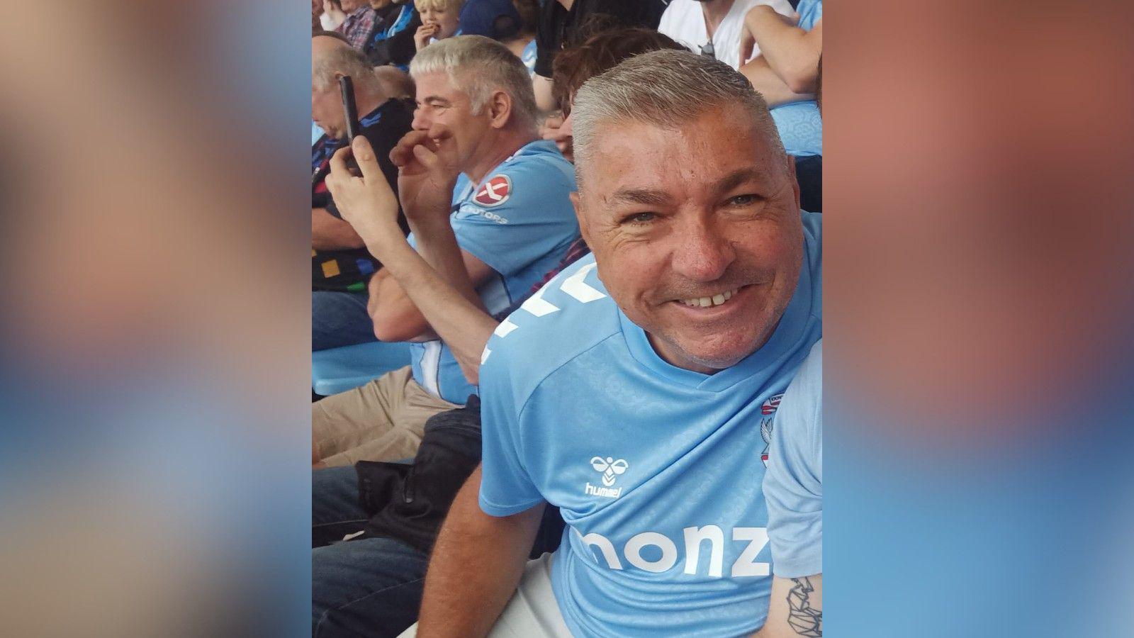 Moz Baker pictured sat down smiling in a light blue Coventry City FC shirt. He has short spiked greying hair.
