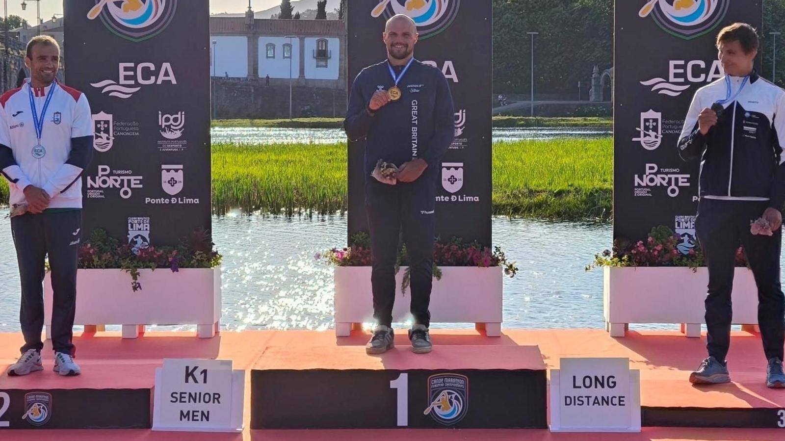 Three men stood on a winners podium.They are smiling and holding bronze, silver and gold medals. the river is behind the podium.