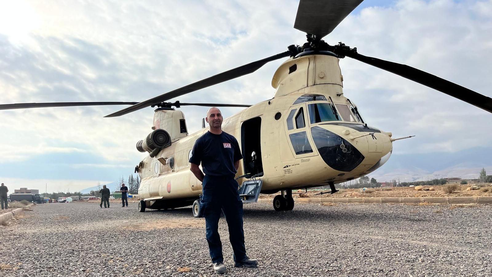 A man stands in front of a helicopter on a broad gravel path. The man man is bald and wearing a black t-shirt and trousers. The helicopter is cream with black blades.