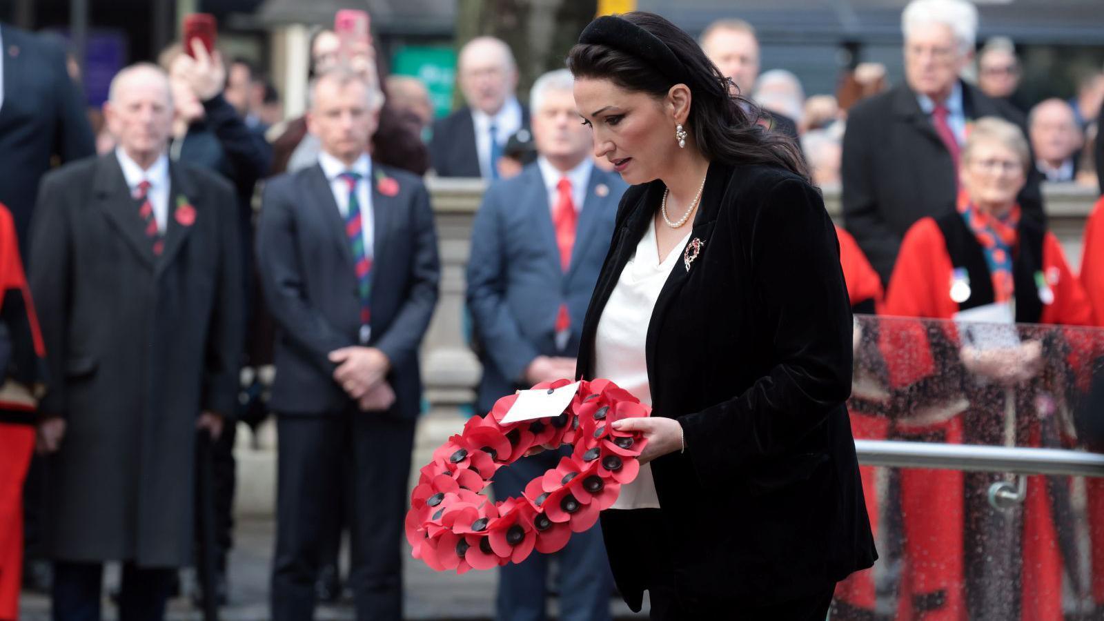 Emma Little Pengelly is holding a poppy wreath. Her head is bent. Crowds of people are watching.