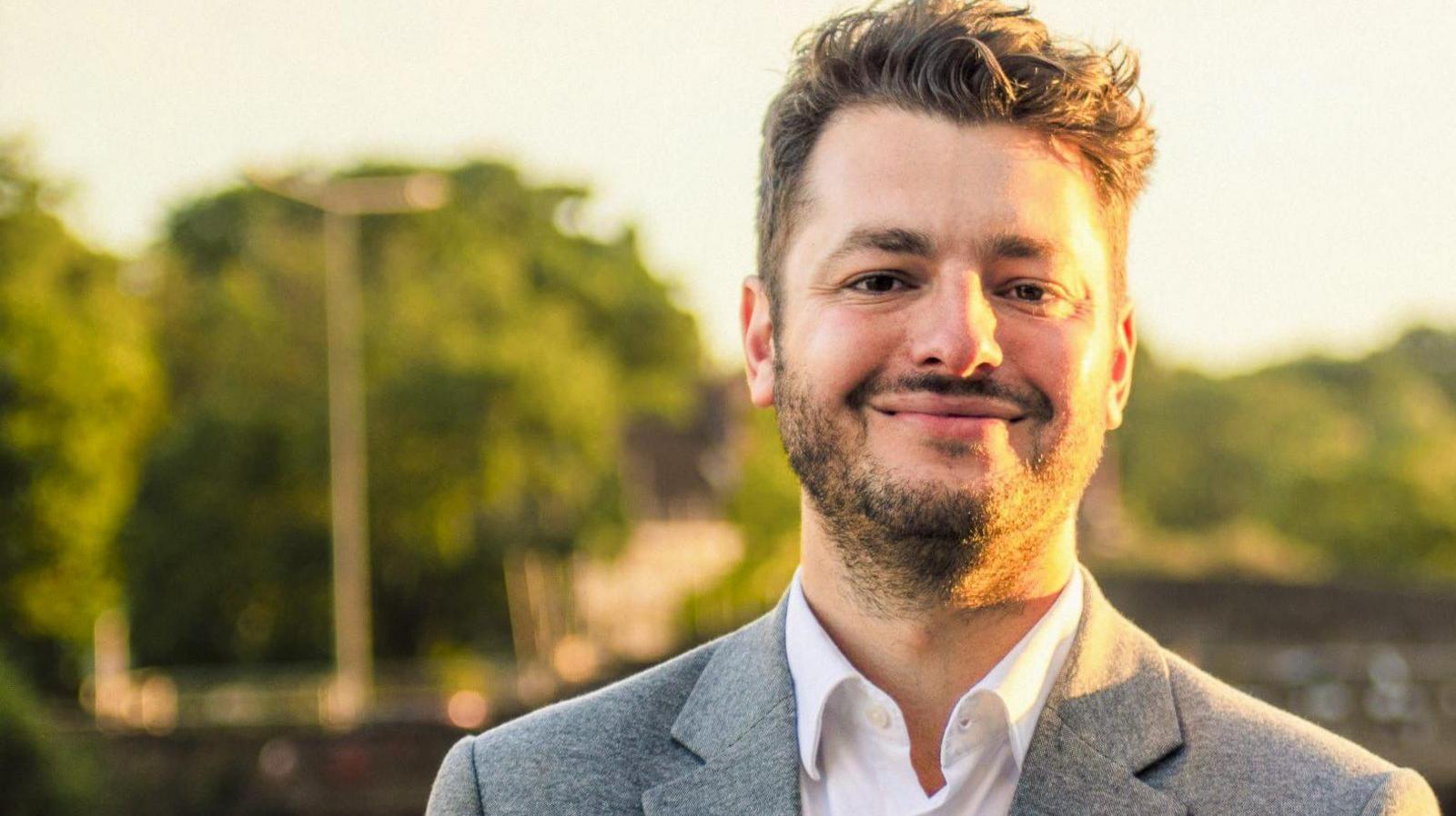 A head-and-shoulders shot of Jack Abbott, who has short brown hair and a short beard and is wearing a white shirt under a grey blazer. He is smiling into the camera.