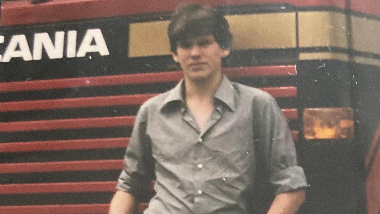 A teenage boy with brown hair wearing a grey long-sleeved shirt rolled up to the elbows, standing in front of a red lorry