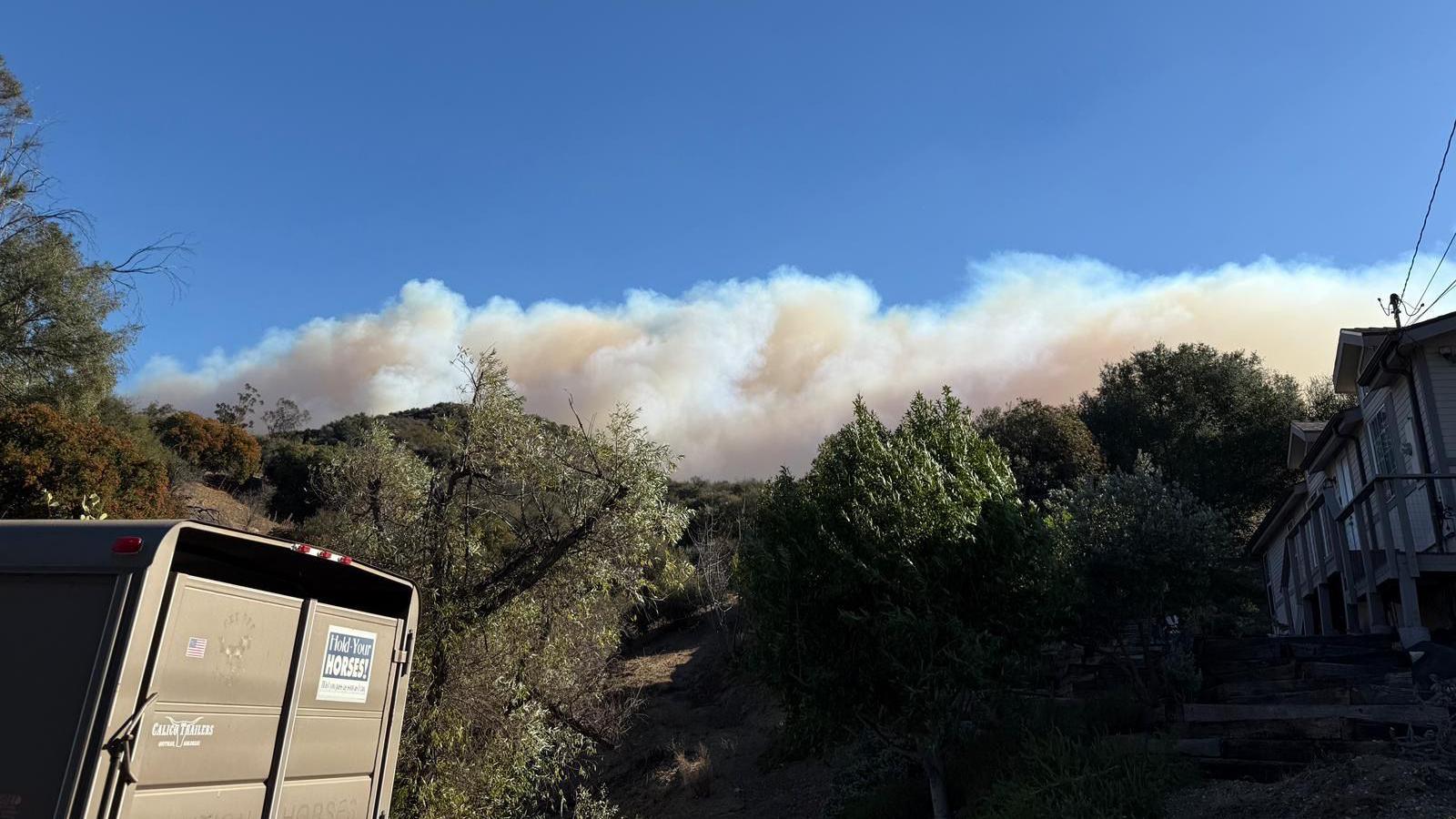 Thick smoke billows above a large hillside against a blue sky. Trees line the hillside. A horse trailer can be seen to the left of the image.