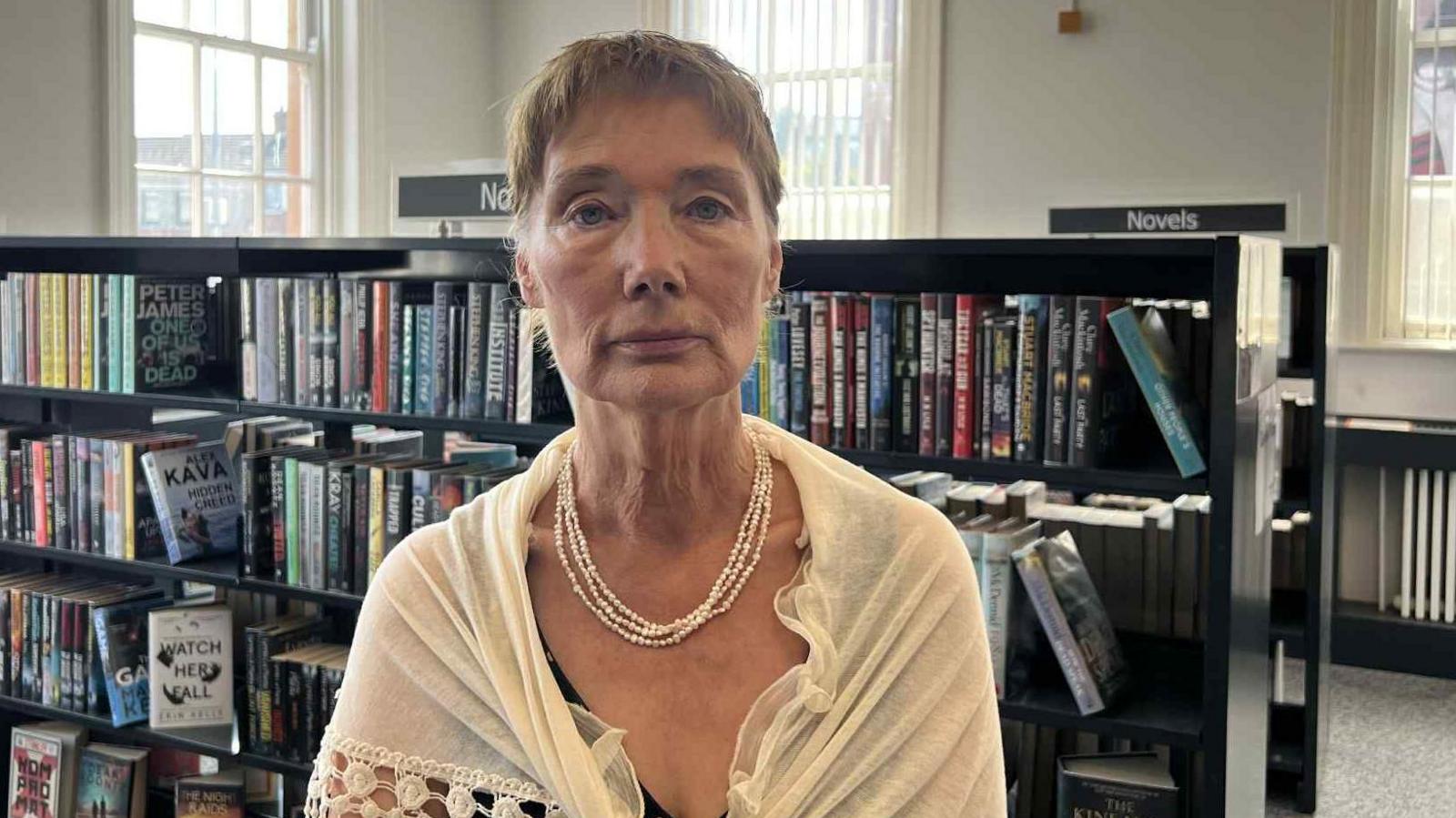 Aine Andrews has short brown hair and is wearing a white shawl and a white necklace. She is standing in the middle of Falls Road Library, black bookcases full of books can be seen behind her.