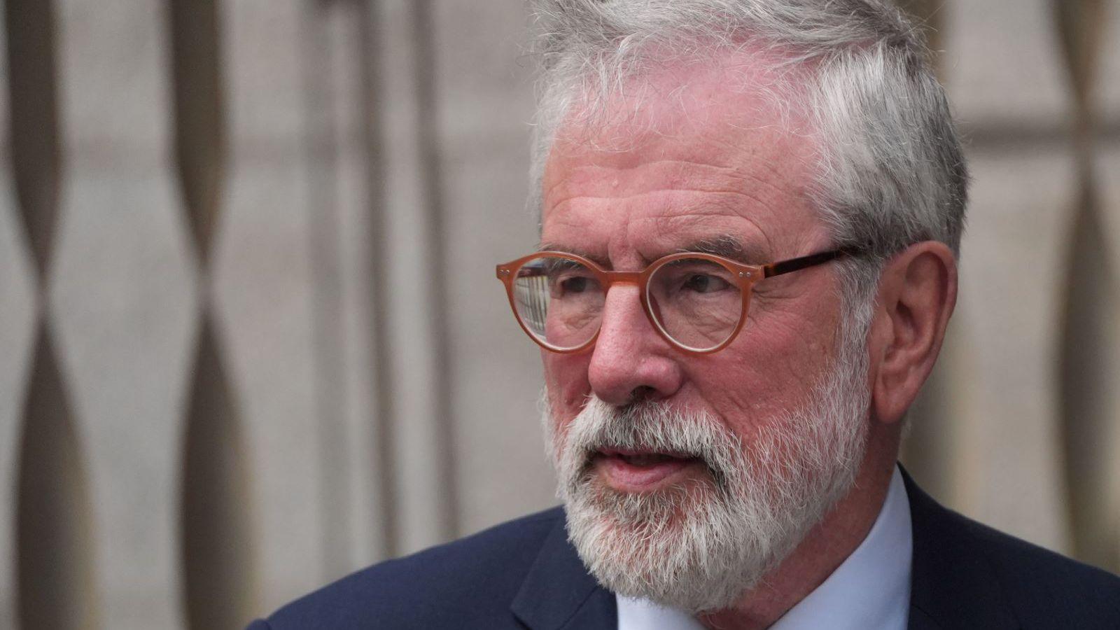 Gerry Adams has grey hair, which is being blown slightly by the wind, and a grey beard. He is looking away from the camera. He has glasses on with orange frames. He is wearing a navy suit jacket and white shirt.  