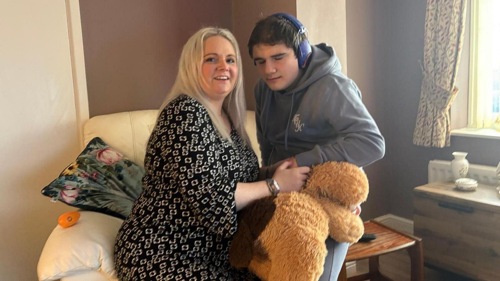 Charlotte and Tobie pictured sitting in a living room. They share a large arm chair. She has a black and white dress on. He is wearing a grey hoodie and holding a large teddy bear. He is wearing blue ear protectors. 