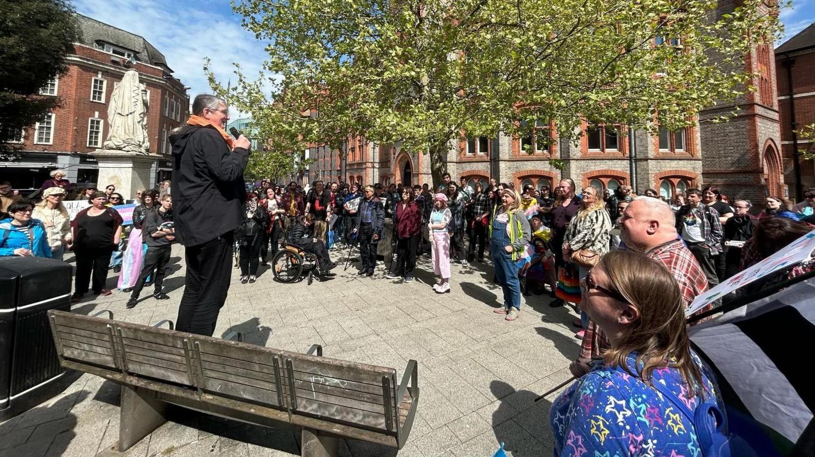 A crowd in a paved square listens to a transgender woman, Kirsten Bayes, who is speaking into a microphone.