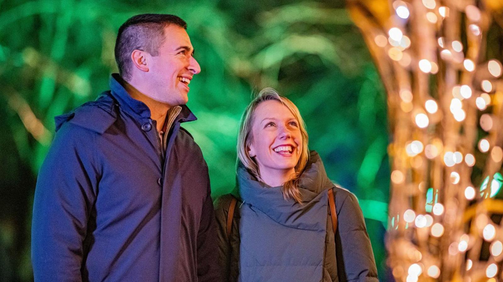 A dark haired man and a blonde woman both wearing dark coats looking up at Christmas lights