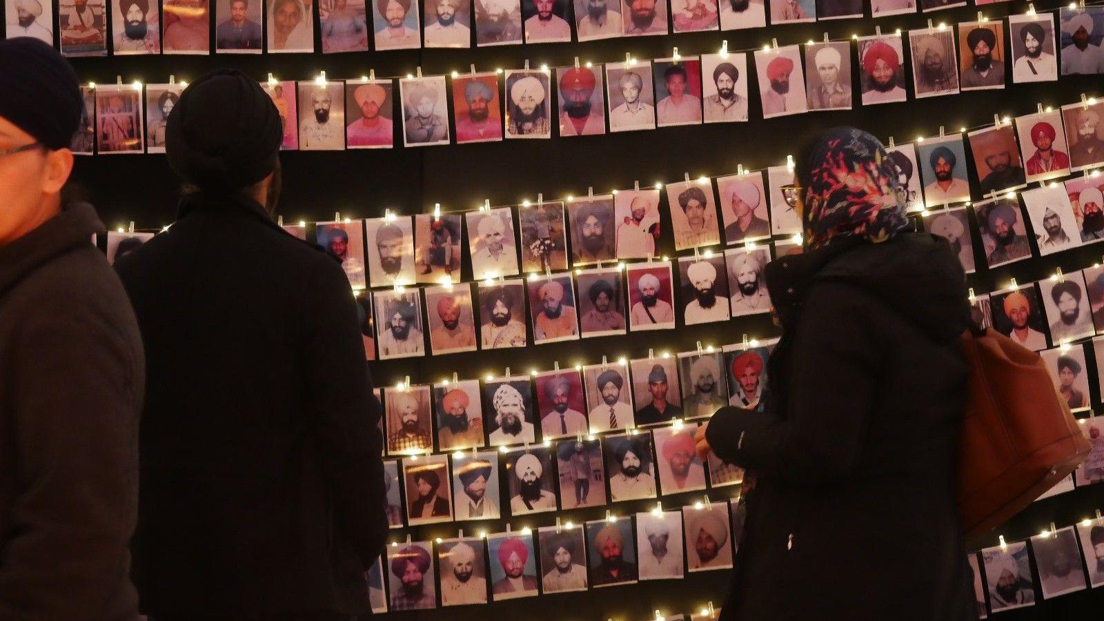 A woman wearing a patterned headdress and black coat, carrying a brown-coloured handbag, and a man wearing a black turban and black coat - both are looking at photographs of people which are hung up in several rows. 